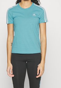 Turkos kortärmad T-shirt med vit Adidas-logga på bröstet och tre vita ränder längs axlarna, tillverkad av mjukt bomullsmaterial.