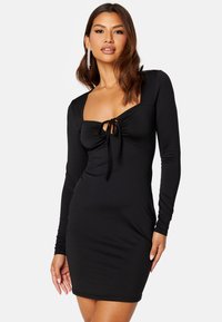 Zwart langmouwig jurkje met een aansluitende silhouet. Heeft een vierkante halslijn en een strikdetail aan de voorkant. Gladde stoftextuur.