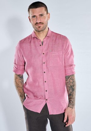 Mann mit kurzen Haaren, der ein pinkes Hemd mit Knöpfen und hochgekrempelten Ärmeln sowie schwarze Hose trägt, zeigt Tätowierungen an den Armen und steht vor hellem Hintergrund.