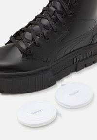 Zapatilla alta negra hecha de material suave, con suela gruesa, cordones texturizados y marca. Dos cordones blancos al lado del zapato.