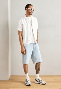 Hvit kortermet linsskjorte over en enkel hvit t-skjorte, satt sammen med lyse denimshorts, hvite ankelsokker og grå New Balance joggesko.