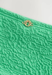 Texturerad mintgrön tyg med ett upphöjt mönster, med en guld cirkulär lås med intrikata detaljer vid den övre kanten.