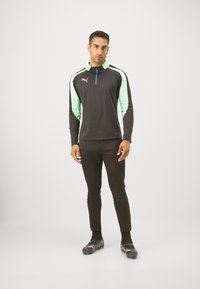 Svart langermet treningsskjorte med mintgrønne ermer og en blå glidelås. Kombinert med svarte bukser og svarte fotballsko.