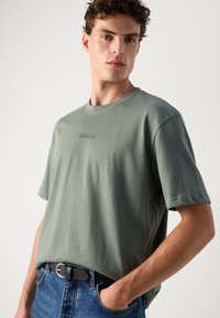 Lys grøn bomulds t-shirt med korte ærmer, rund halsudskæring og tonalt "BOSS" logo i midten; parret med blå denimjeans og et sort bælte.