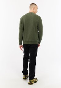 Groene sweatshirt met lange mouwen en een ronde halslijn, gecombineerd met zwarte broek en multikleure sportschoenen. De trui heeft een klein logo detail.