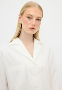 White, button-up shirt with a pocket and a subtle logo. Smooth texture, classic collar, and relaxed fit. Model with long straight hair and earrings.