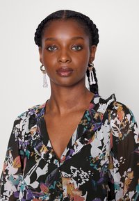 Blusa com padrão floral em preto, com colarinho franzido, apresentando cores vibrantes como roxo e azul-turquesa. Modelo usa brincos com contas penduradas.