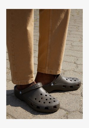 Des Crocs noires avec des trous de ventilation, une surface texturée et une sangle arrière, associées à un pantalon marron clair, sur une surface pavée.