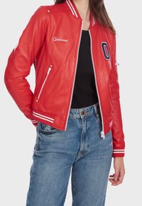 Kobieta w czerwonej skórzanej kurtce Oakwood z białym zamkiem i paskami przy mankietach, w połączeniu z niebieskimi dżinsami i czarną bluzką.