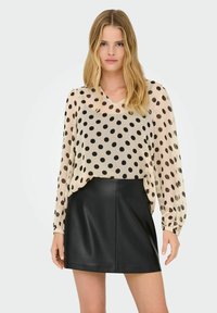 Gennemsigtig beige bluse med sorte prikker, lange ærmer og v-hals, parret med en sort læder miniskirt. Glat tekstur og tætsiddende design.