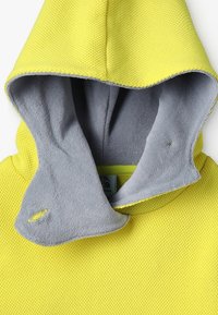 Žlutý kapucňový oděv s texturovaným povrchem a měkkým šedým podšívem. Disponuje klokaní kapsou a jemnými prvky stehu.