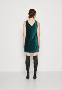 Tummansininen samettinen mini mekko V-mallisella selällä, yhdistettynä reiteen yltävään mustaan saappaan. Sujuva tekstuuri, hihaton muotoilu ja yksinkertainen siluetti.