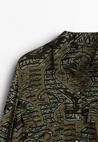 Long-sleeve shirt in dark olive green with abstract black and textured white patterns, displaying a buttoned placket and collar.