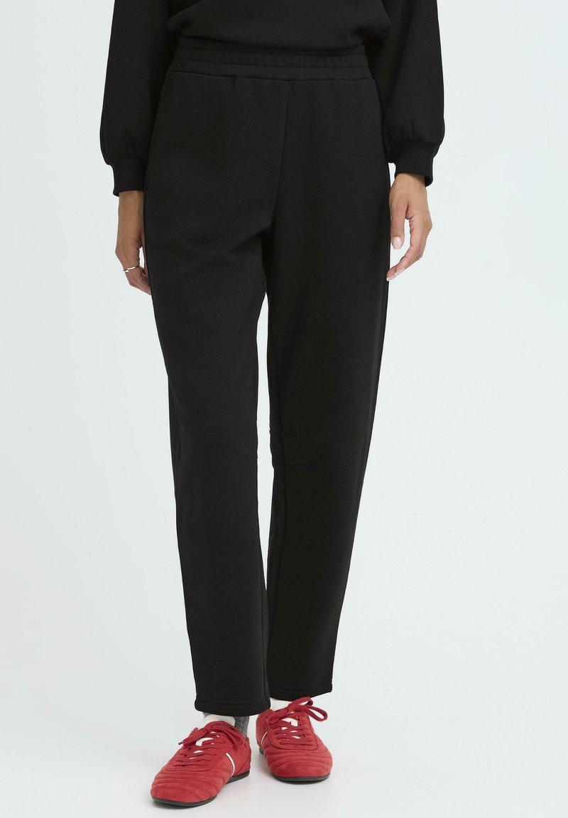 Melnas sporta bikses, kas izgatavotas no mīksta auduma, ar elastīgu jostu, divām sānu kabatām un šauru kāju dizainu. Apvienotas ar sarkaniem apaviem.