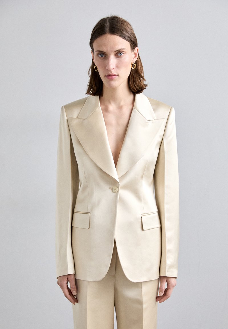 Beige satinblazer med djup V-ringning, strukturerade axlar, två framfickor och enkel knäppning. Slät textur och skräddarsydd passform.