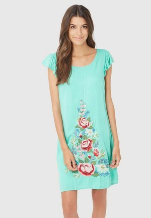 Joven con cabello largo y castaño que lleva un vestido turquesa con mangas acampanadas y un gran bordado floral en la parte delantera.