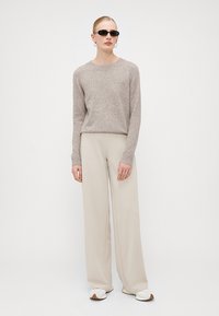 Maglione lavorato a maglia beige con collo a giro, abbinato a pantaloni beige a gamba larga. Il modello indossa occhiali da sole scuri e sneakers bianche. Design semplice e texturizzato.
