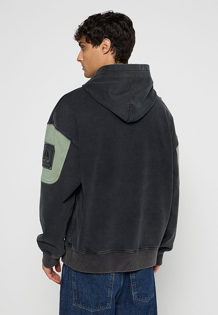 Kapuzensweatshirt aus dunkelgrauem Baumwollmix mit lockerer Passform, das ein grünes Akzent-Patch am linken Ärmel und einen gerippten Saum aufweist.