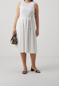 Robe blanche sans manches avec un bustier texturé et une ceinture en corde contrastante, associée à un sac en satin marron et des sandales à talons noires.