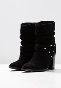 Musta mokka nilkkuri, jossa on löysä muotoilu, paksu korko ja soljettiedot hopeisilla yksityiskohdilla. Sujuva pinta.