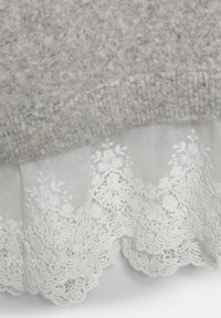 Szara wełniana tkanina nakłada się na białą koronkę wykończoną haftem w kwiaty, z falbanym brzegiem. Tekstura kontrastuje pomiędzy materiałami.