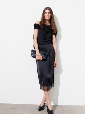 Femme en top noir épaules dénudées et jupe midi en satin avec ourlet en dentelle, tenant une pochette noire, debout contre un mur blanc.