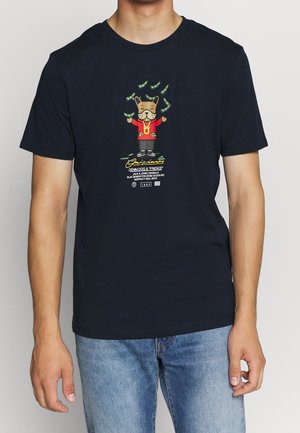 Mężczyzna w granatowym T-shircie z kreskówkowym psem w czerwonej kurtce, złotym łańcuszkiem i unoszącymi się wokół dolarami, w połączeniu z jasnoniebieskimi dżinsami.
