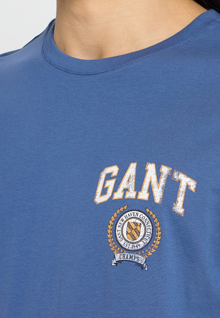Blaues Baumwoll-T-Shirt mit rundem Ausschnitt, das ein weißes und goldenes "GANT"-Grafikdesign sowie ein kreisförmiges Logo mit Lorbeerakzenten auf der Brust zeigt.
