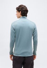 Άνδρας με την πλάτη, φοράει ανοιχτό γαλάζιο υφασμάτινο πουκάμισο μακρυμάνικο με ψηλό γιακά και μαύρο παντελόνι cargo, μπροστά σε απλό φόντο.