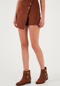 Shorts en daim marron avec un design enroulé, accentués par deux boutons. Associés à des bottes mi-mollet marron avec quincaillerie décorative et une sangle.