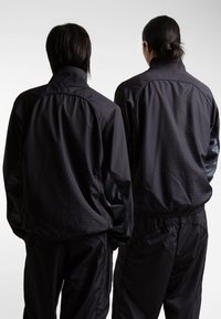 Two individuals wearing matching black track jackets and pants. The jackets feature a subtle geometric pattern and stand-up collars.