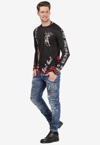 Schwarzes Langarmshirt mit Tigermotiv und weißem Text, kombiniert mit blauen Cargo-Jeans mit mehreren Taschen und Used-Look, schwarze Stiefel.