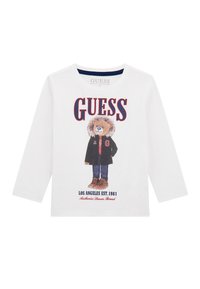 T-shirt bianca a maniche lunghe realizzata in cotone organico, con una grafica di un orso in giacca e stivali marroni. Il testo include "GUESS" e "LOS ANGELES EST. 1981."