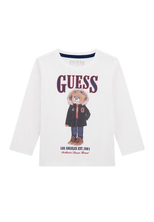 T-shirt bianca a maniche lunghe realizzata in cotone organico, con una grafica di un orso in giacca e stivali marroni. Il testo include "GUESS" e "LOS ANGELES EST. 1981."
