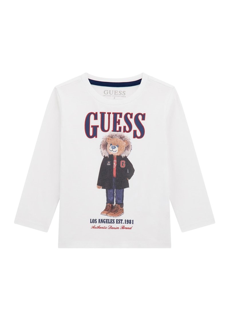 T-shirt bianca a maniche lunghe realizzata in cotone organico, con una grafica di un orso in giacca e stivali marroni. Il testo include "GUESS" e "LOS ANGELES EST. 1981."