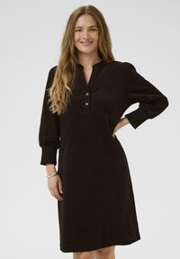Bruine corduroy jurk met lange pofmouwen, een kraaghalslijn en knoopdetails aan de voorkant. Gladde textuur en knie-lengte ontwerp.
