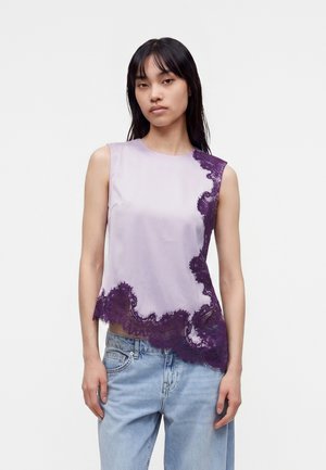 Femme portant un haut lavande sans manches avec des détails en dentelle asymétriques violet foncé et un jean bleu clair, debout devant un fond uni.
