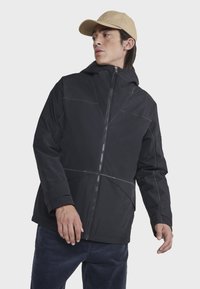 Veste à capuche noire avec des coutures d'accent grises, fermeture éclair à l'avant et coupe décontractée. Le modèle est associé à un pantalon en velours côtelé bleu marine et à une casquette beige.
