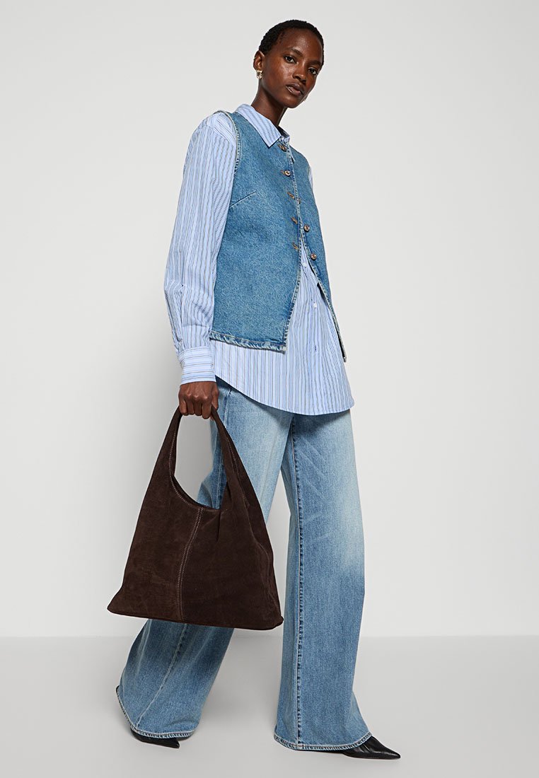 Chaleco de mezclilla sobre una camisa a rayas azul y blanca, combinado con unos vaqueros de pierna ancha de color azul claro y un bolso de hombro de ante marrón. Zapatos negros de punta.