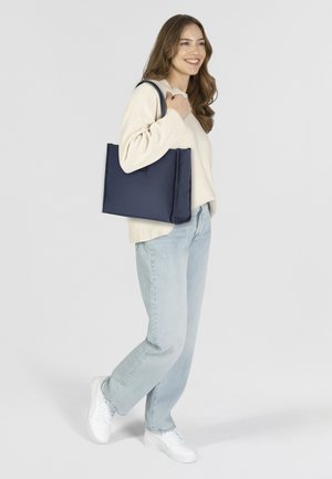 Borsa a spalla blu navy con design morbido e imbottito. Presenta manici robusti e forma rettangolare. Indossata a tracolla con denim casual.