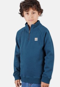 Blå fleece pullover med kvart lynlås ved halsen, raglanærmer og ribbede manchetter. Lille firkantet logo-patch på venstre bryst.