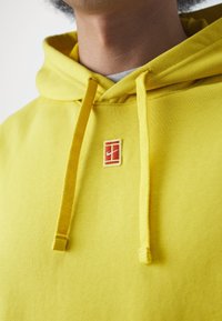Sweat à capuche jaune en tissu doux ; comporte un petit écusson logo rouge et blanc ainsi que des cordons assortis. Design simple avec une coupe classique.