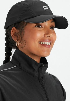 Schwarze Kappe mit einem gebogenen Schirm, ausgestattet mit einem reflektierenden Logo und Stitching-Details. Kombiniert mit einer schwarzen Reißverschlussjacke und Hängerohrringen.