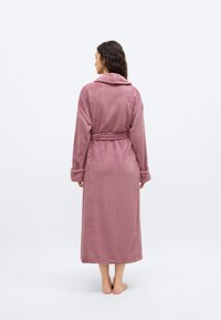 Γυναίκα που στέκεται ξυπόλητη με την πλάτη, φορώντας ένα μωβ απαλό μπουρνούζι μέχρι τον αστράγαλο, δεμένο στη μέση, με φόντο ένα απλό λευκό φόντο.