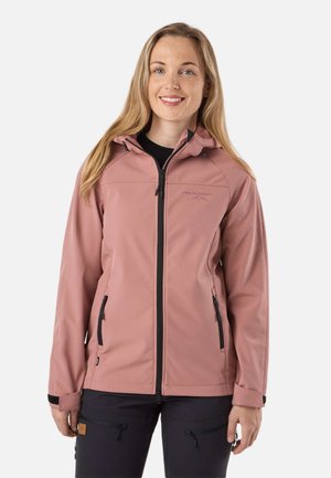 Femme portant une veste zippée rose poudré avec des poches zippées noires et un pantalon foncé avec des poches zippées, debout devant un fond uni.