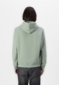 Felpa verde chiaro con cappuccio regolabile, texture morbida, polsini e orlo a costine; abbinata a jeans grigi scuri, mostrando un design semplice e casual.