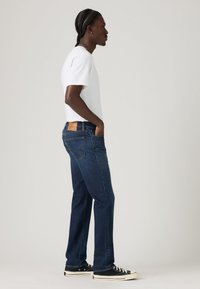 Jeans de denim azul oscuro con un corte recto, con bolsillos traseros y un parche de cuero en la cintura. Llevados con una camiseta blanca y zapatos negros.