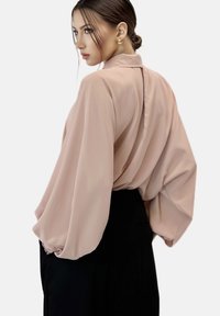 Blusa rosa chiaro con maniche oversize, realizzata in tessuto morbido, presenta un collo alto e un dettaglio a chiave sul retro. Abbinata a pantaloni neri.