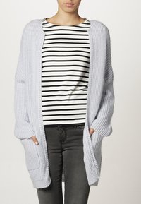 Hellgraue Strickjacke mit offenem Vorderteil, gerippter Textur und zwei vorderen Taschen, getragen über einem schwarz-weißen gestreiften Oberteil.