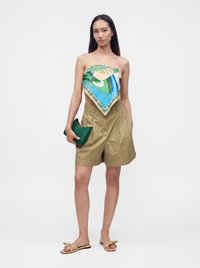 Femme portant un haut foulard coloré sans bretelles, un short taille haute kaki, des sandales beige avec un nœud, tenant une petite pochette verte.